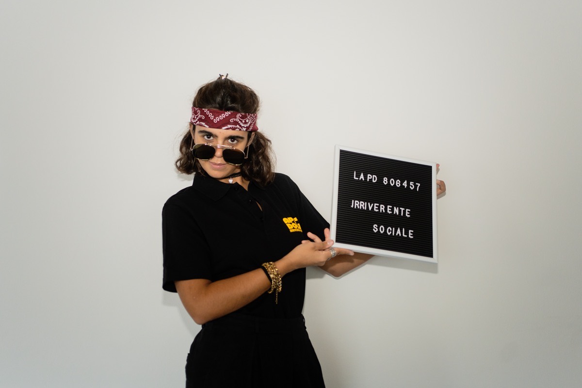 Una ragazza con bandana e occhiali da sole, polo nera Ape'n Down, indica un cartello con la scritta Irriverente sociale