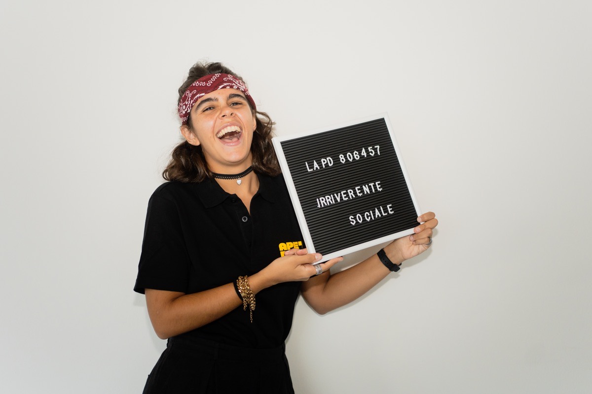 Una ragazza con la polo nera Ape'n Down ride e mostra un cartello con la scritta Irriverente sociale